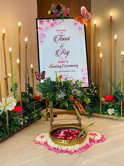 A beautiful welcome sign for a baby's naming ceremony. The decor features a floral and butterfly theme, with elegant candle-like lights and a traditional brass bowl of rose petals.