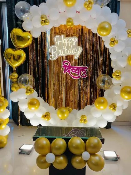 A classic first birthday decoration in white and gold. This setup includes a full balloon ring, a shimmer backdrop, a personalized name sign, and a number '1' balloon column.