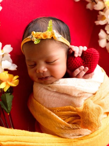 A sweetly smiling newborn holds a red knit heart, swaddled in a yellow wrap and surrounded by yellow and white flowers.