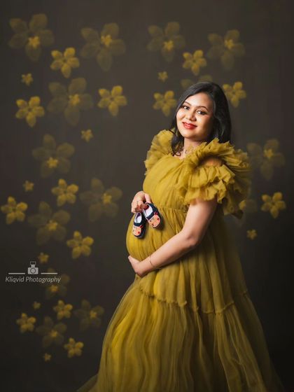 Holding tiny shoes against her bump, this mother-to-be glows in a ruffled olive green gown against a floral backdrop.