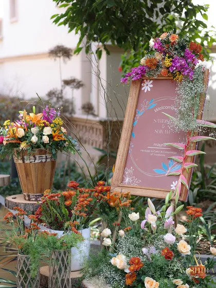 A welcome sign for the 'Tropical Tales' event, framed with a beautiful arrangement of colorful flowers in a wooden bucket.