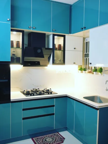 A bold and vibrant modular kitchen design. The high-gloss turquoise and white acrylic cabinets create a fresh and energetic atmosphere, complemented by a marble-patterned backsplash and smart storage solutions like a dedicated spice rack.