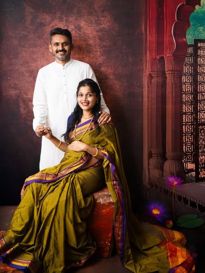 An elegant portrait of an expecting couple in traditional attire. The rich olive green saree and classic backdrop evoke the feeling of a timeless Indian painting.