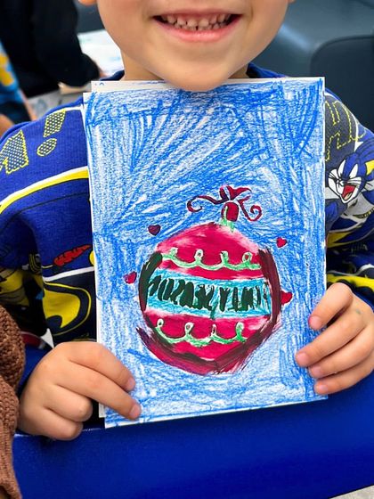 A proud student holding up his hand-drawn Christmas card, complete with a colorful ornament.
