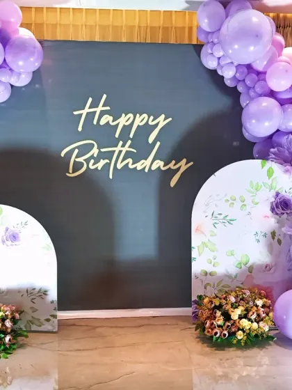 A beautiful lavender-themed birthday decoration. This setup uses a large grey backdrop with smaller floral-printed arches, all tied together with a lovely purple balloon garland.