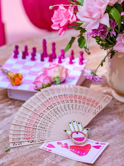 A playful composition of a custom playing card, a beaded Hamsa hand brooch, and a vintage fan, with the pink chessboard in the background. These details make an event truly personal.