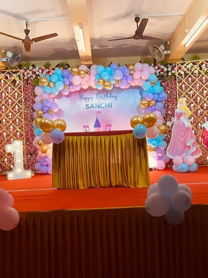 A dreamy Disney Princess themed stage for a first birthday. The decoration features cutouts of various princesses like Ariel and Cinderella, a pastel balloon arch, and a backdrop with a castle silhouette.