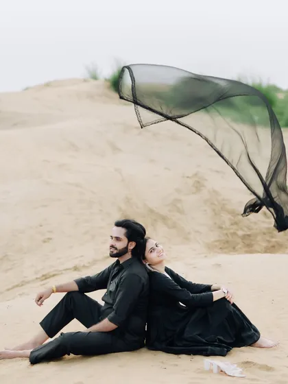 A serene moment in the sand dunes. The couple sits back-to-back while a black dupatta catches the wind, adding a touch of graceful movement to this peaceful portrait.