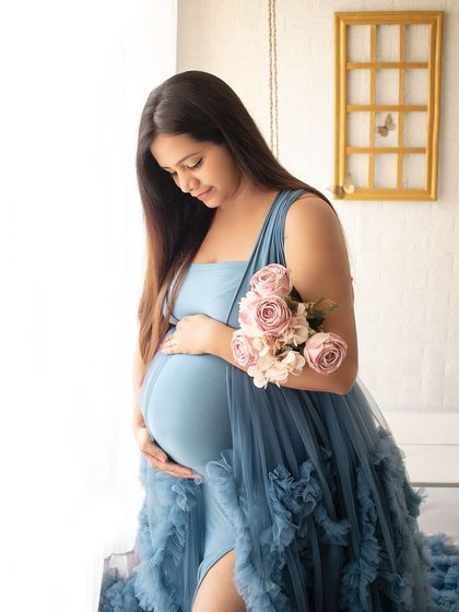 A close-up solo portrait of a mother-to-be in a blue gown, holding a bouquet of pink roses and looking lovingly at her bump.