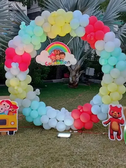 An outdoor Cocomelon themed party with a full circular balloon arch in rainbow colors and character cutouts.