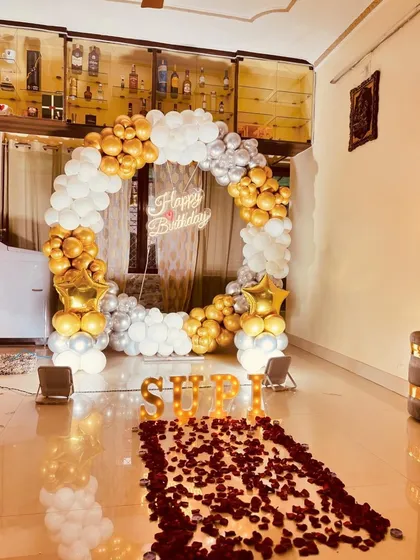 A personalized birthday surprise set up in a living room. This decoration features a gold and white balloon arch, a path of rose petals, and the name 'SHRI' in illuminated marquee letters for a custom touch.
