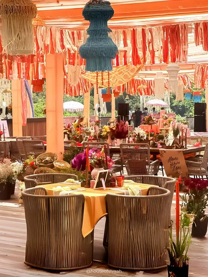 A dining table at the Pahadi Mehendi, with rustic rope-woven chairs and a colorful tassel chandelier hanging overhead, creating a vibrant and textured look.