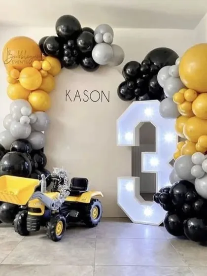 A construction-themed party featuring a toy truck, a large "3" marquee number, and a balloon arch in yellow, black, and grey.