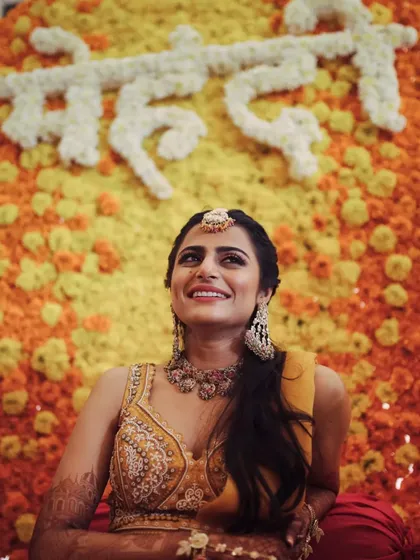 A joyful, upward-looking shot of the bride, with the word 'Mehendi' written in white flowers on the marigold wall.
