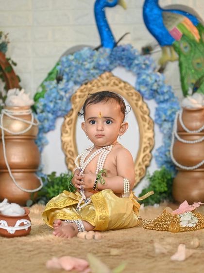 A thoughtful Little Krishna, dressed in yellow with peacock-themed decor. Each Krishna shoot is unique, with different props and color schemes.
