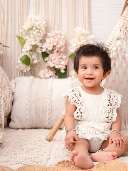 A happy baby girl in a lace romper, perfectly matching the boho-chic aesthetic of the macrame and wicker props.