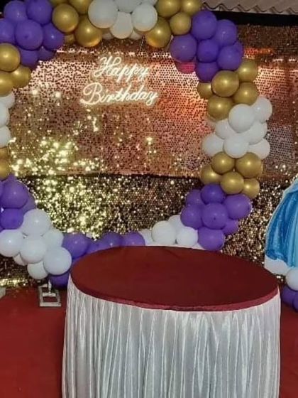 A glamorous birthday backdrop with a gold sequin wall and a purple, white, and gold balloon arch. The 'Happy Birthday' neon sign adds a modern, trendy touch.