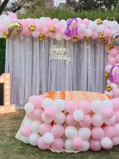 A beautiful first birthday party with a Disney Princess theme, set up outdoors. This decor includes a pink and white balloon arch, a sheer curtain backdrop with fairy lights, and a light-up number '1'.