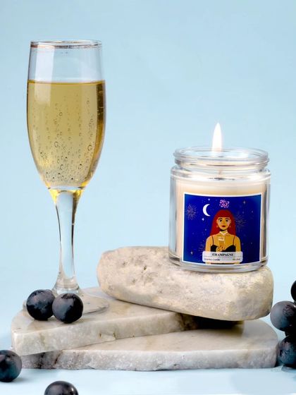 Here, the champagne candle rests on natural stone props next to a bunch of dark grapes and a flute of champagne. This composition blends earthy textures with celebratory elegance.