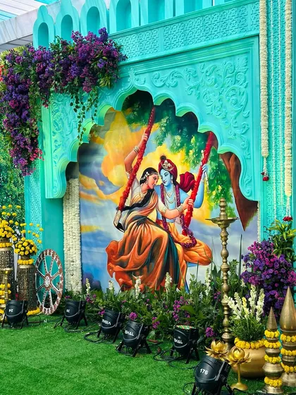 This is a centerpiece from a Vrindavan-themed event, featuring a beautiful custom-painted backdrop of Radha and Krishna on a swing. The setup is framed by intricately carved panels and lush floral arrangements, bringing a divine and artistic feel to the celebration.