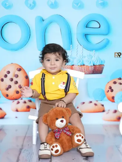 A baby boy in a yellow shirt sits with his teddy bear in a cookie-themed first birthday setup. The giant cookie props and blue background create a fun and unique celebration.