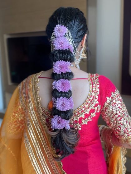 A vibrant and beautiful hairstyle for a Haldi ceremony. This intricate bubble braid is woven with fresh purple flowers, adding a pop of color that complements the festive outfit.