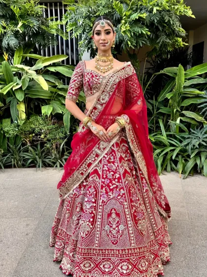 A full view of a model in a traditional red bridal lehenga, with her hair in a floral bun. The updo is positioned to perfectly accommodate a matha patti and dupatta.