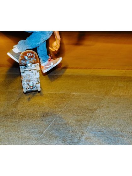An overhead action shot of a skater's feet on their board. The motion blur and warm lighting create a dynamic and gritty feel, typical of street skate photography.