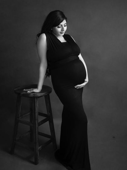 A full-length solo portrait in black and white. The mother-to-be leans against a stool, creating a simple yet elegant composition.