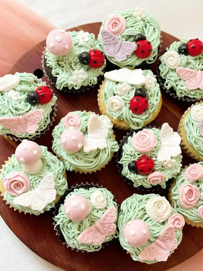 A dozen enchanted garden cupcakes with light green buttercream piped to look like grass. The set is decorated with delicate fondant details, including pink butterflies, red ladybugs, pink mushrooms, and small flowers for a whimsical woodland theme.