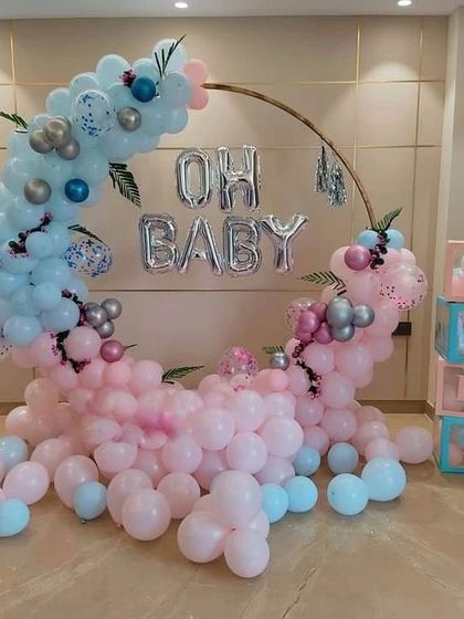 An "Oh Baby" gender reveal setup with a crescent-shaped balloon arrangement on a ring frame. The design uses pink and blue balloons, "BABY" blocks, and silver foil letter balloons.