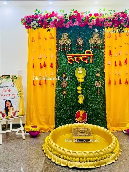 A festive Haldi setup with a green backdrop, yellow drapes, and traditional hangings. The design includes a welcome sign and a central urli for the ceremony.
