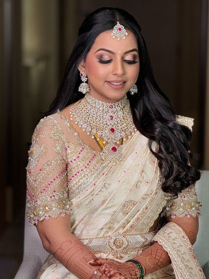 A beautiful bride in a white and gold saree with a sheer, embellished blouse. The makeup is glamorous, with a focus on defined eyes and glowing skin.