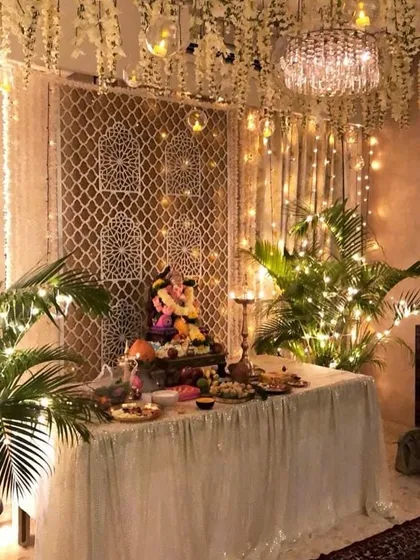 A beautiful bohemian-style Ganpati decoration. The setup features a macrame backdrop, hanging wisteria, potted palms, and lots of fairy lights, creating a warm and intricate look.