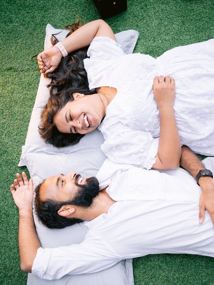 A fun and relaxed moment from a pre-wedding shoot. A couple shares a laugh while lying on the green lawn, showcasing a casual and intimate style of photography.