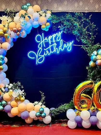 A vibrant circular balloon arch for a 60th birthday, decorated with greenery and a bright blue neon sign.