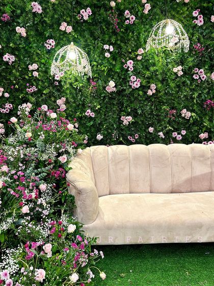 A beautiful engagement stage with a lush green flower wall dotted with pink roses. The elegant couch and delicate hanging chandeliers add a touch of sophistication and romance to the setting.