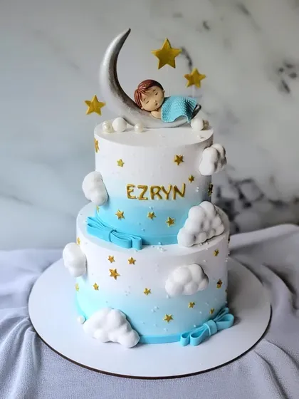 A sweet christening cake for a baby boy. This two-tier cake in blue and white is decorated with clouds, stars, and a baby sleeping on the moon.