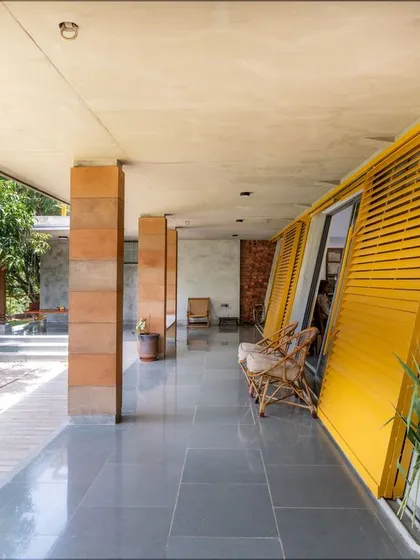 The deep verandah, or 'Jagli', runs along the house, defined by its inclined walls and bright yellow louvered shutters. This semi-open space provides a shaded transition between the interior and the lush green landscape.