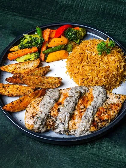 A full meal platter with grilled chicken in a creamy sauce, rice, potato wedges, and sautéed vegetables. The dark green background provides a rich, earthy tone to the composition.