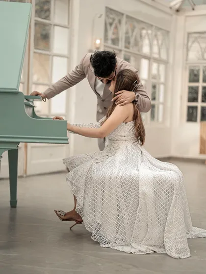 A passionate and romantic moment at the piano. The groom leans in to kiss his bride's shoulder as she plays, creating a deeply intimate and cinematic photograph.