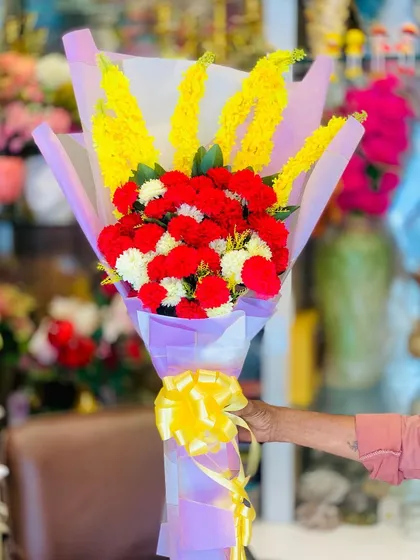A bright and cheerful artificial flower bouquet. This arrangement features red and white carnations with tall yellow accent flowers, wrapped in purple paper.