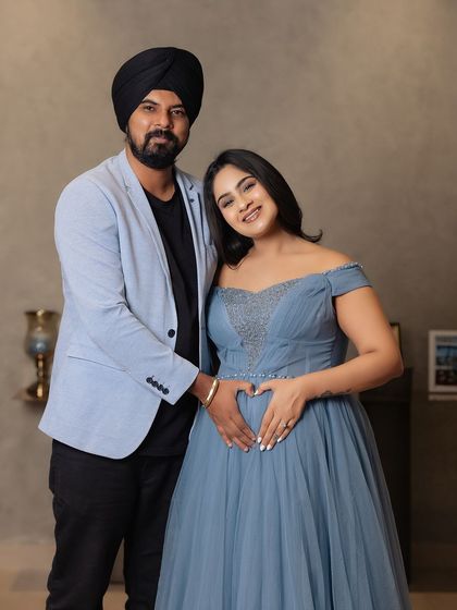 A joyful and classic couple portrait. They stand together, smiling, with their hands forming a heart on the baby bump, celebrating their growing family.