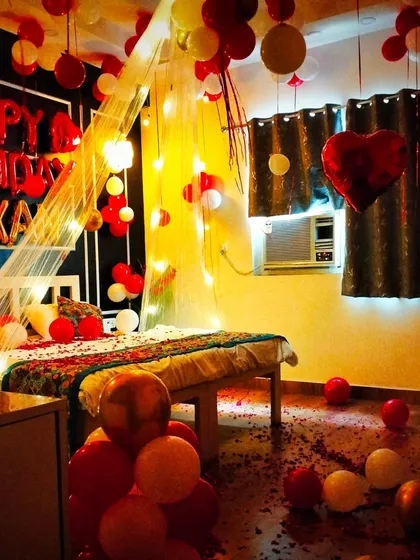 This room decoration has a cozy and warm feel, with yellow lighting complementing the red and white balloons. The canopy and scattered petals make it perfect for a couple's celebration.