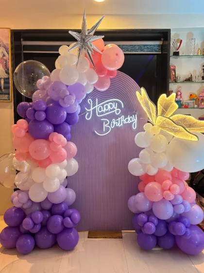A clear, well-lit shot of the butterfly-themed birthday backdrop. The design features a pastel purple arch, a neon sign, and two columns of organic balloons in shades of purple, pink, and white, accented with a star foil balloon.