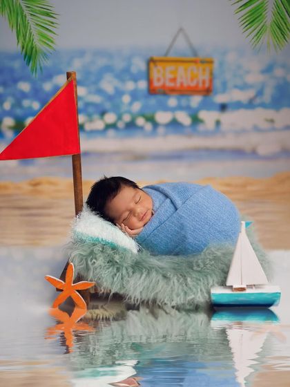 A creative beach-themed newborn shoot. The baby is sleeping soundly by the "water," complete with a miniature sailboat and a beach backdrop.