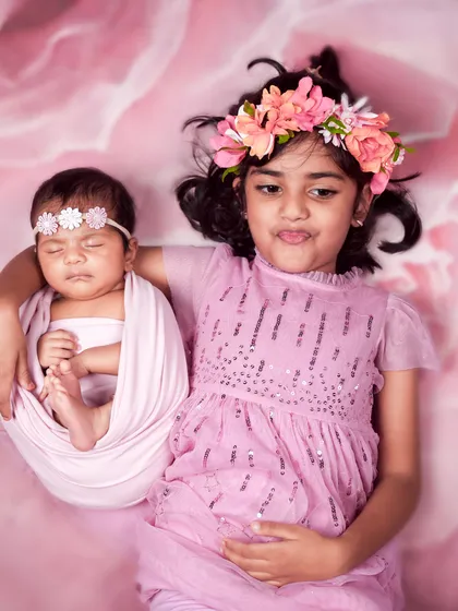 An older sister holds her new baby sibling in a sling-like wrap against a floral pink backdrop. This creative pose is a fun way to include siblings in the shoot.