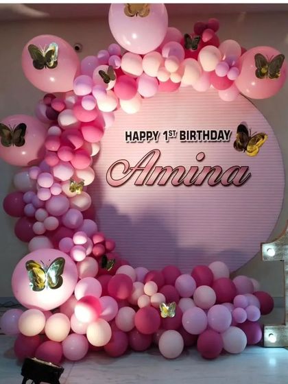 A beautiful butterfly-themed first birthday in shades of pink. The decor features a personalized backdrop with the name 'Amina', a full balloon arch, and a light-up number '1'.