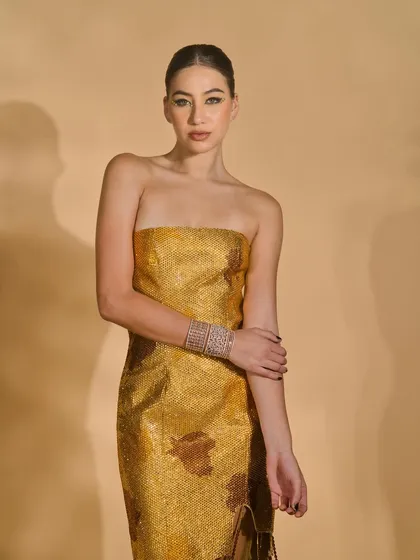 A medium shot of a model in a strapless gold textured dress from the 'Ivory Edit'. The lighting is carefully controlled to emphasize the unique, shimmering texture of the fabric.