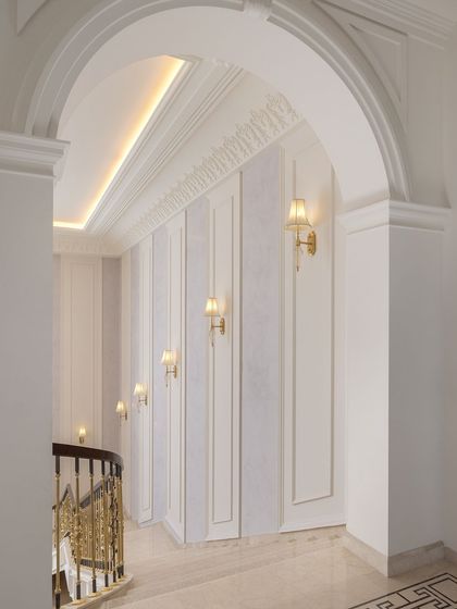 This corridor in The Ivory Royale celebrates Indian craftsmanship and classical design. The arched entryway, ornate wall sconces, and detailed balustrade lead the eye towards the grand staircase, enhancing the overall aesthetic of the space.